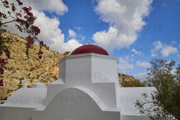 A white church with a red dome, surrounded by plants and rocks under a dramatic blue sky with