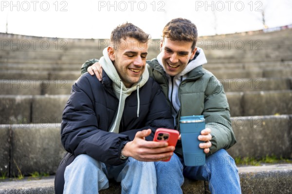 Two men are sitting on steps, embracing and looking at a smartphone while one holds a reusable