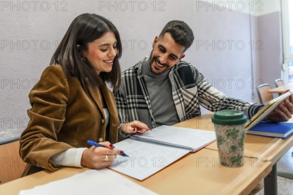 Two happy university students are studying together in a classroom, taking notes and using a