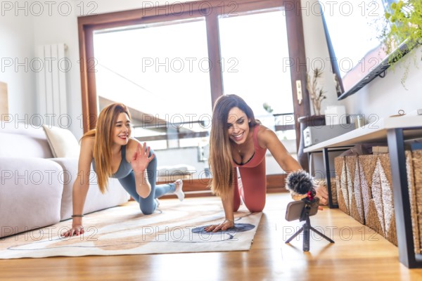 Two women in sportswear smiling and recording an online yoga class in their modern living room,