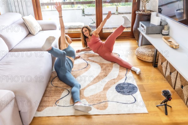 Two smiling women lying on carpet practicing yoga together at home following online class on
