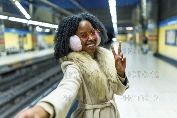 Smiling woman wearing earmuffs and pink glasses making selfie and victory sign while waiting subway