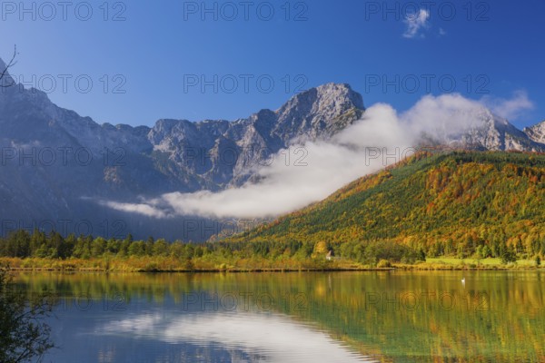 Lake Almsee in autumn with a forest reflected in the lake. A slowly dissipating fog moves over the