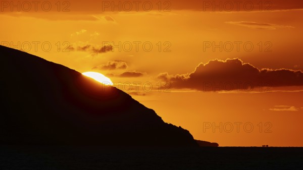 Soft orange light of the sun behind a mountain, cloud formations in the sky, sunset, Kassos,