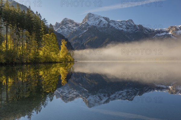 Lake Almsee in autumn with a forest and mountains reflected in the lake. A slowly dissipating fog