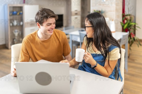 Young multiethnic couple smiling and looking at each other while using laptop and drinking coffee,
