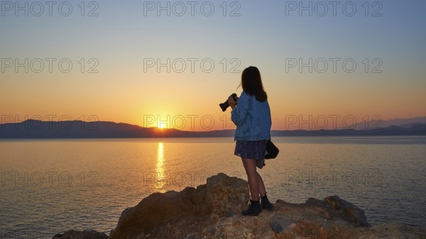 Sunrise, sun reflection on the sea, orange-blue cloudless sky, girl from behind with jeans jacket
