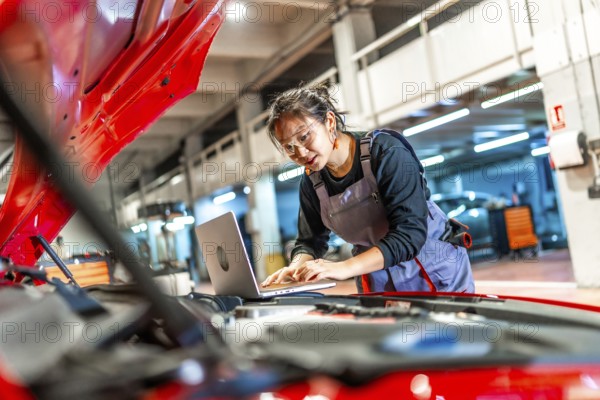 Young female mechanic working on a laptop while inspecting a car engine in a modern workshop,
