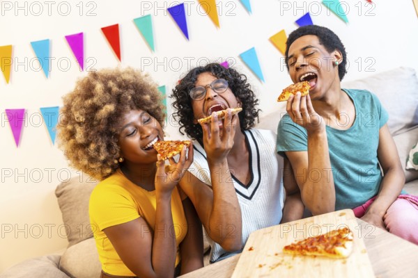 Multi-ethnic friends biting a slice of pizza having fun sitting on the sofa together at home