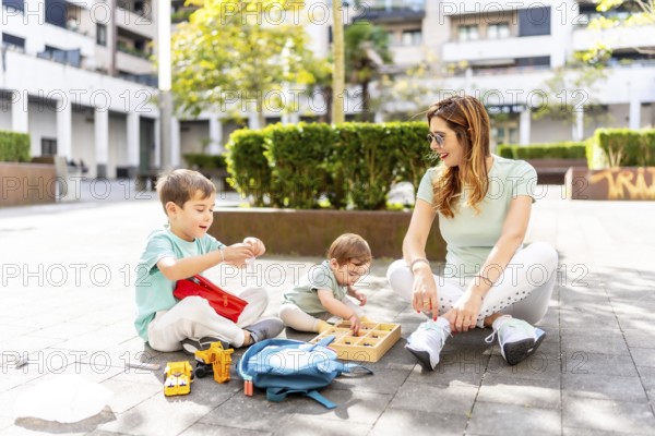 Full length photo of a caucasian mother and brothers sitting on urban park playing with toys