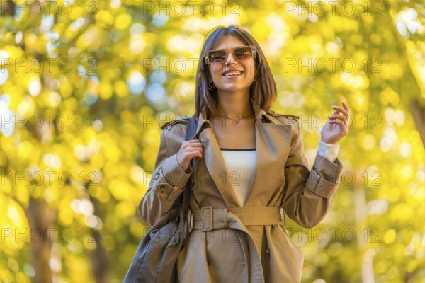 A woman wearing a tan coat and sunglasses is walking down a path. She is smiling and holding a