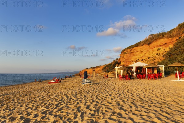 Beach café, red rocks illuminated by the evening sun, sandy beach with tourists, holiday by the