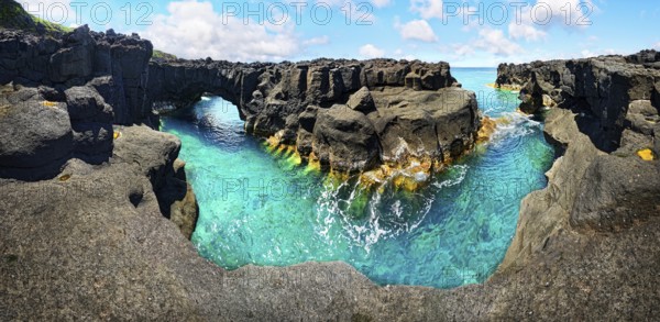 Panoramic view of rugged lava rock formations with a natural arch over turquoise sea under blue