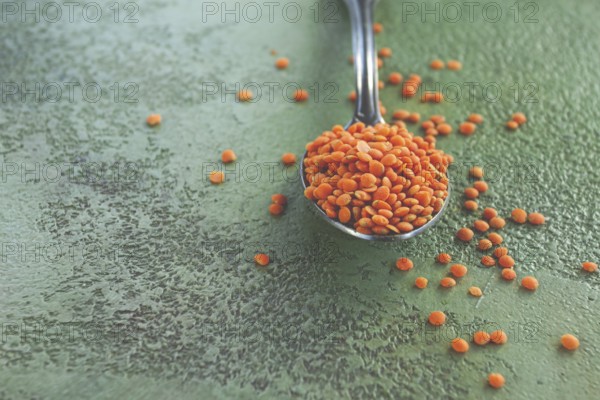 A spoon holds vibrant red lentils, with more lentils scattered around on a textured green surface.