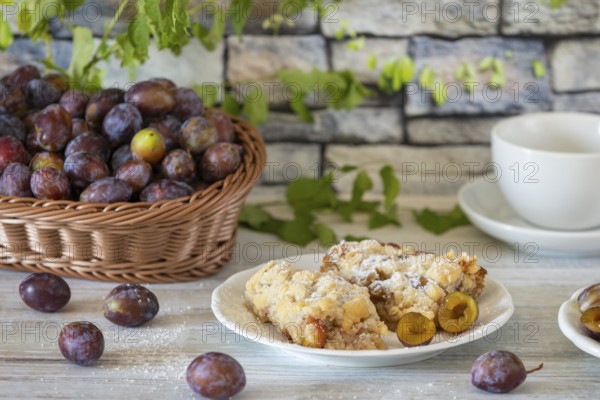 Plum cake on a plate next to a basket with fresh plums and a cup in the background, on a white