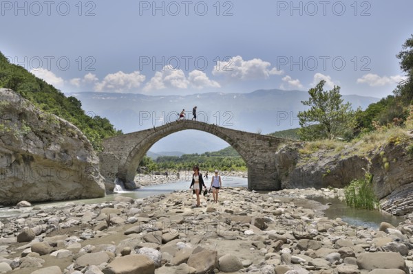 Two young woman walking barefoot in the riverbed over large stones, Ottoman stone bridge, Kadiut