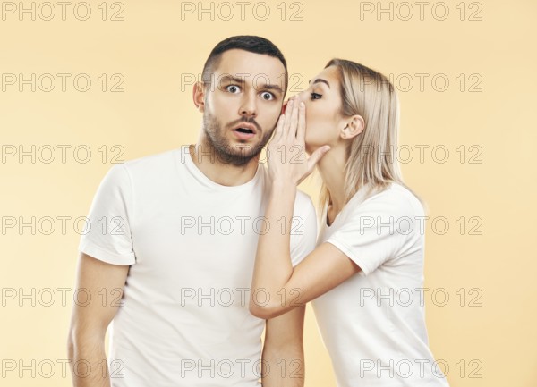 Young woman whispering secrets to man over beige background. Gossiping, couple relationship concept