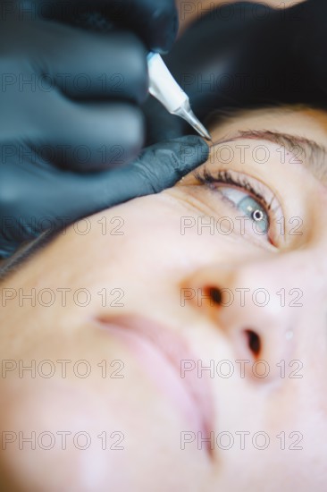 Beautician applies permanent makeup to a woman's eyebrow in a busy salon, showcasing precision and