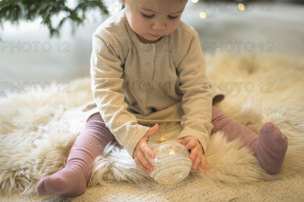 Toddler sitting on a rug and looking at a Christmas tree bauble in a cosy setting