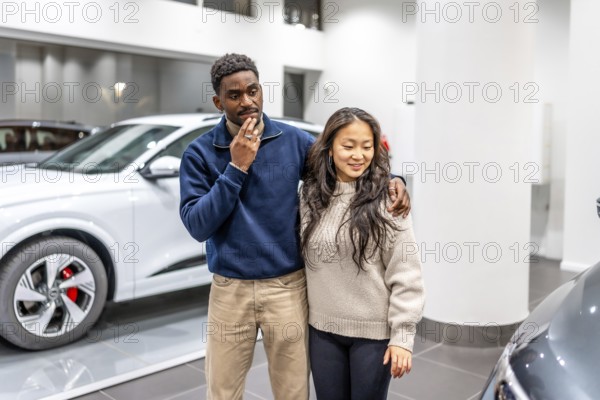 Happy multiethnic couple choosing their new electric car in a dealership showroom, sustainable