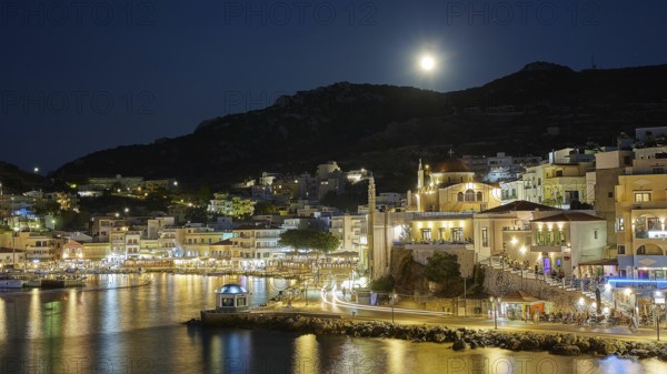 Illuminated coastal town at night with moon in the sky and water in the foreground, Rising full
