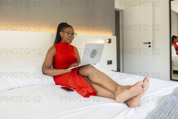 Businesswoman wearing a red dress using a laptop while sitting comfortably on a hotel bed during a