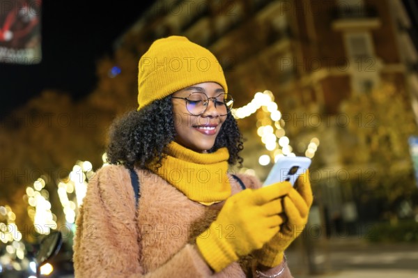 Smiling young woman wearing yellow winter clothes using smartphone in a city street at night with