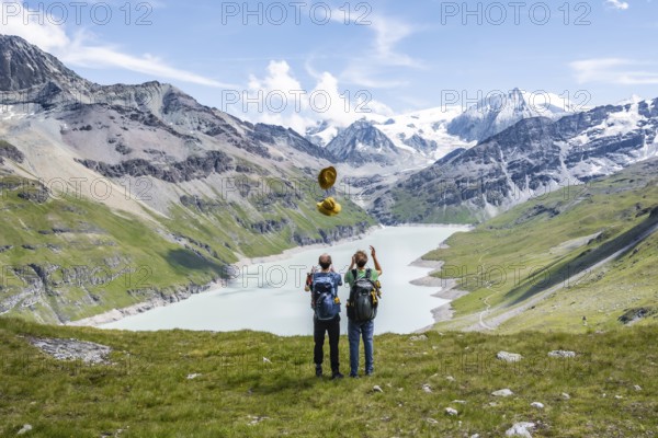 Two friends throwing yellow hats in the air, mountaineers in front of a blue mountain lake, Lac des