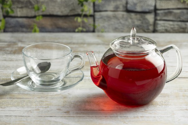 A teapot with red tea next to an empty glass cup with spoon on a table in front of a stone