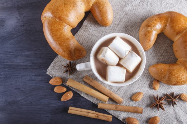 A cup of hot chocolate with marshmallow, croissants and spices on linen napkin and black wooden