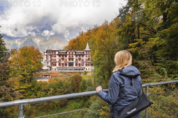 A woman looks from a railing at a large hotel surrounded by autumnal forest, Lake Brienz, Giessbach