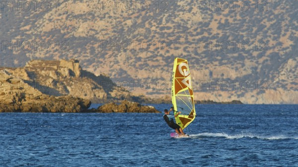 Person windsurfing in front of rocky coastal scenery and blue mountains in the background,