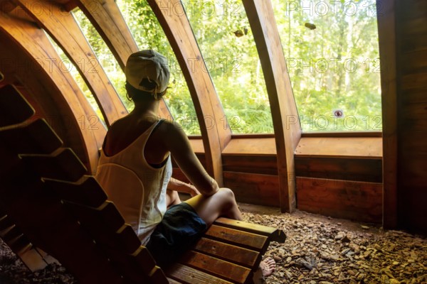 A woman is sitting on a wooden bench in a forest. She is wearing a white tank top and blue shorts.
