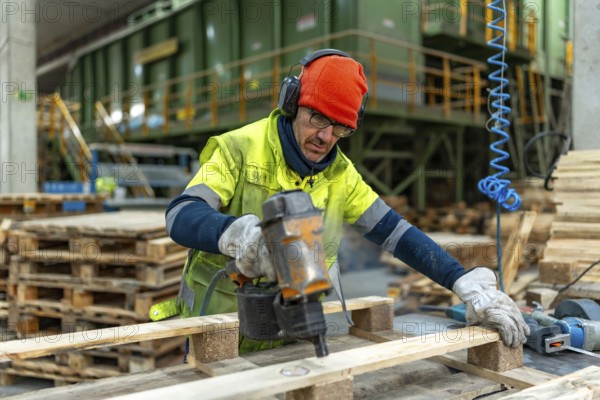 Man with protective gear repairing pallets in a recycling plant warehouse using a compressed air