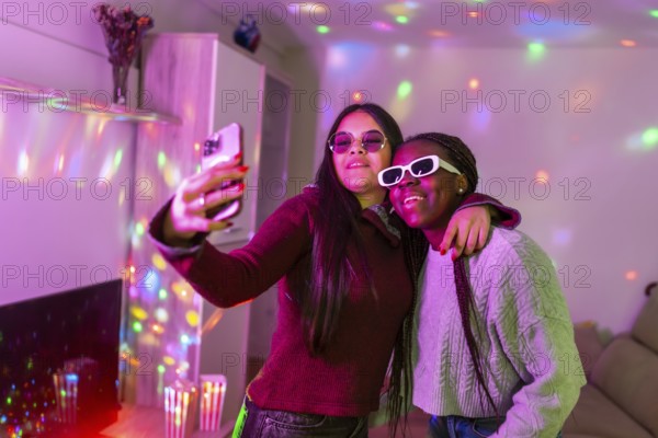 Two teenage girls wearing sunglasses are taking a selfie with a smartphone at a home party with