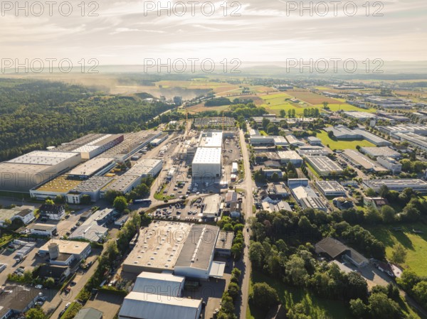 Numerous buildings and streets in an industrial area, surrounded by wooded landscape under a sunny