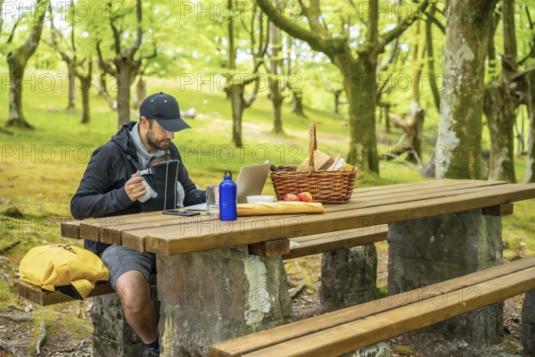 A young Caucasian father teleworking sitting at a picnic table with computer in the woods. Summer