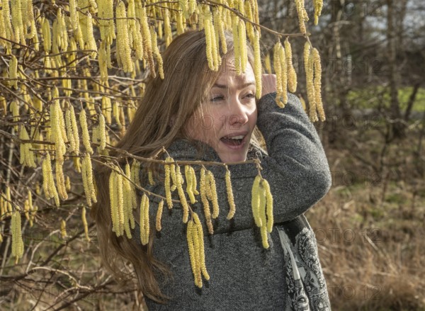 Young Caucasian woman with allergy problem stands by flowering hazel bush and sneezes in the crook