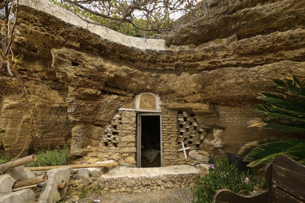 Exterior view of a cave carved into the rock with a stone wall and vegetation around the entrance,