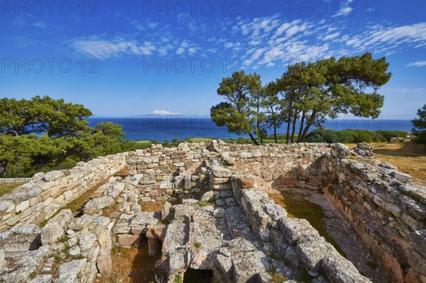 Ancient ruins with stone walls and views of the sea and trees under a blue sky, Kamiros,