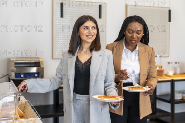 Two businesswomen are enjoying breakfast at a hotel buffet, adding jam to their toast during a