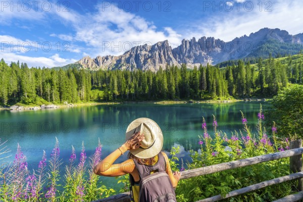 Woman with backpack and hat admiring the turquoise waters of lake carezza with the latemar mountain
