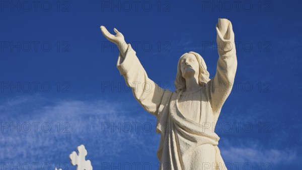 A stone statue of Jesus with outstretched arms against a blue sky, Agios Nikolaos church, cemetery,