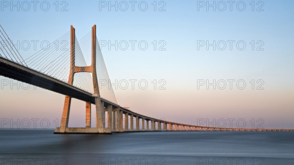 A long cable-stayed bridge spans over water under a gradient sunset sky, Lissabon, Vasco Da
