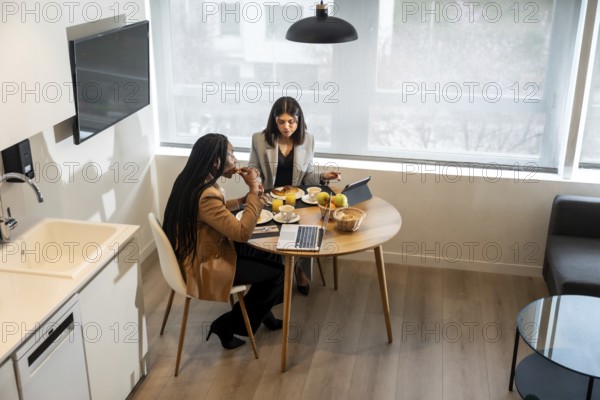 Two businesswomen are having breakfast in their hotel room while working on a tablet and laptop