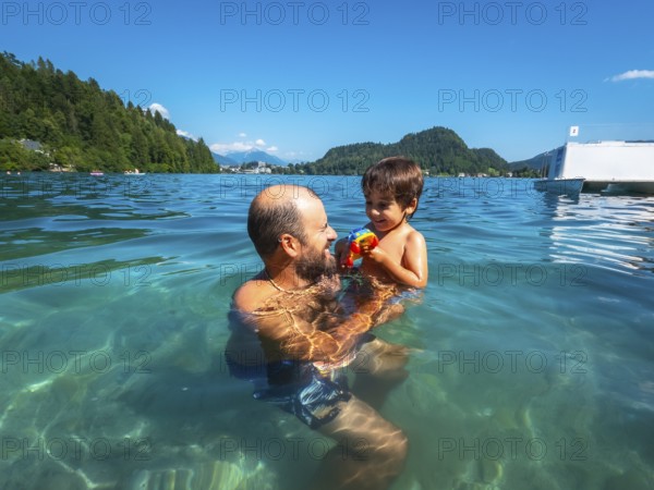 Father playing with his son in the clear water of lake bled, with mountains and forest in the