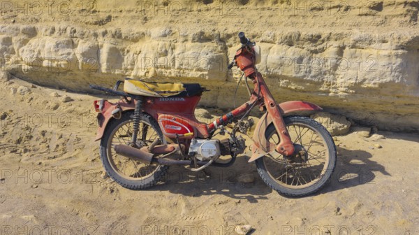 An old Honda C50 motorbike stands abandoned in front of a rocky cliff, Paralia Agios Theodoros