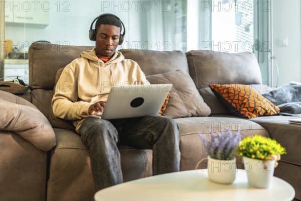 Young man concentrating on his laptop while wearing headphones, comfortably seated on a sofa in his