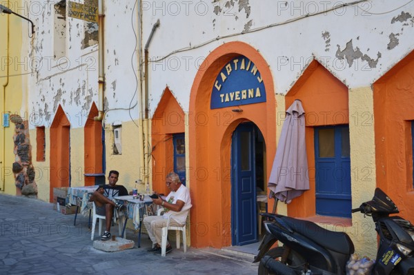 People sitting in front of a colourful facade of a taverna and enjoying their time on the street,