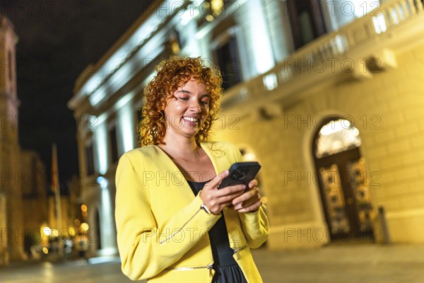 Smiling cheerful woman in yellow clothes and curly hair using smartphone walking along city at